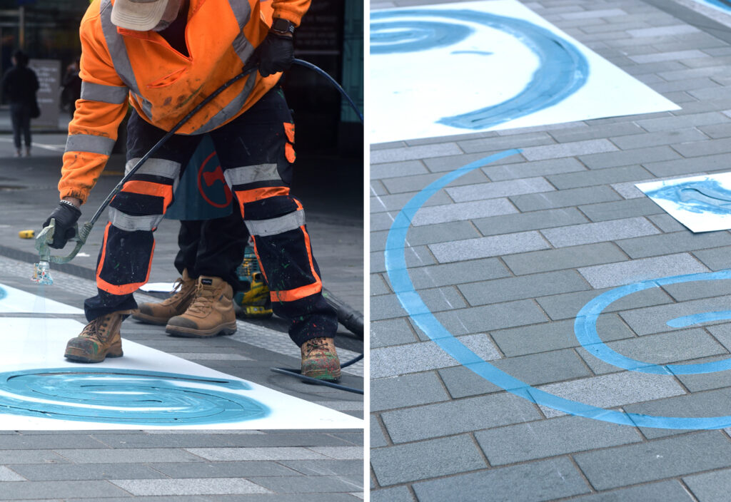 Two images of the Waihorotiu Path design being applied to the sidewalk. Image one of a painter painting on the stencil. Image two is a detail showing the stencil and the design on the sidewalk.